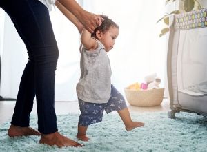 Enfant en short bleu et t-shirt gris essayant de faire ses premiers pas avec sa maman qui lui tient les mains.