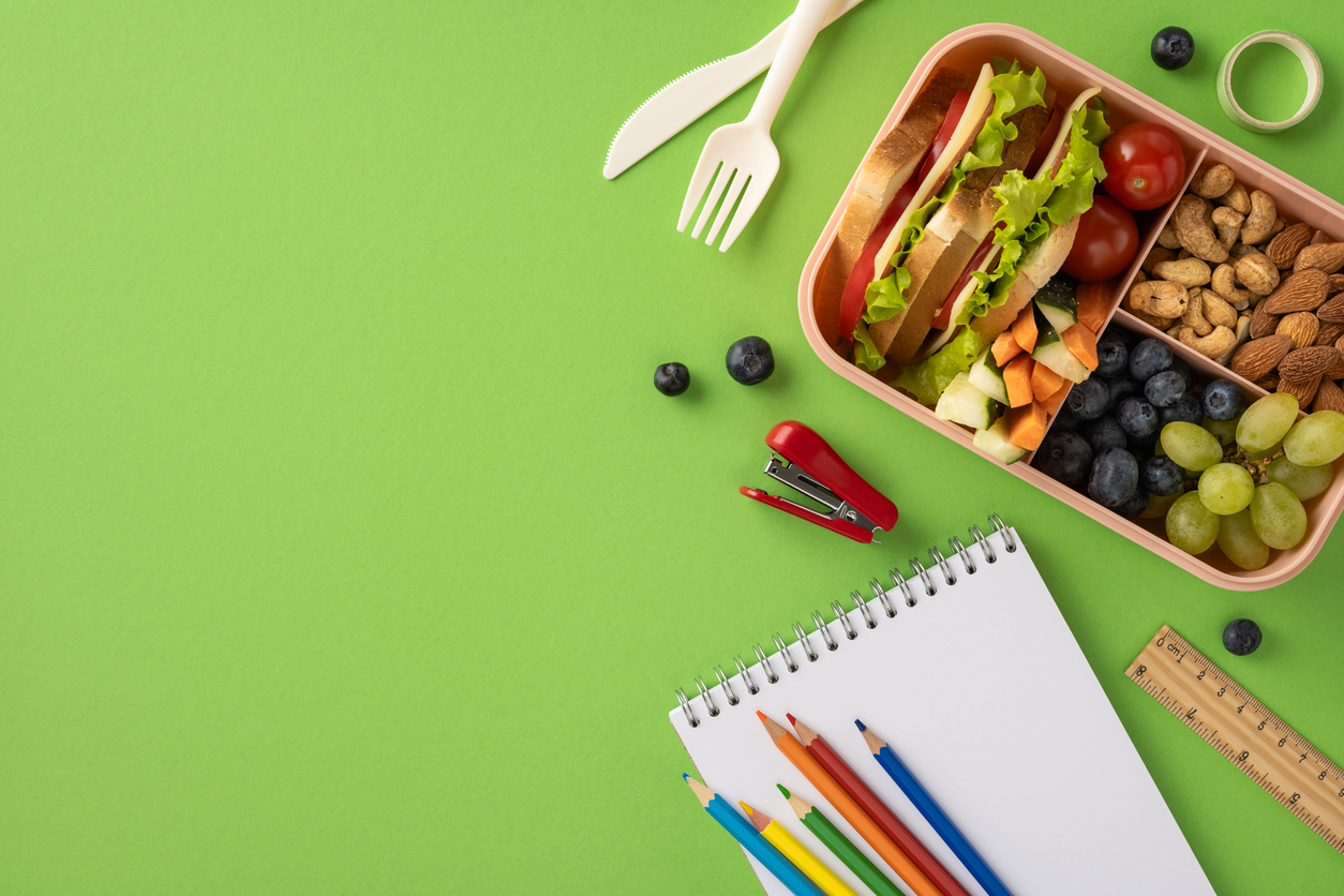 A prepared lunchbox containing nutritious food items alongside school supplies arranged on a green surface